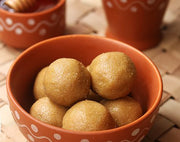 Traditional millet snacks, shaped into balls and placed in a terracotta bowl, with a decorative pattern on the side. The snacks appear to be sweet, coated with a glistening ingredient, possibly jaggery.